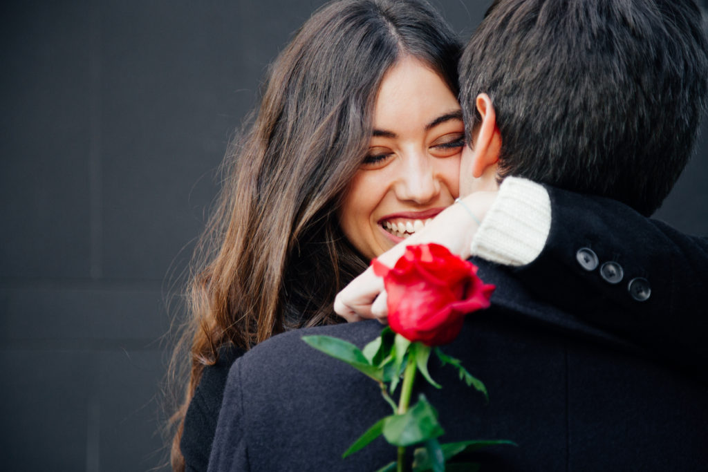 Beautiful and happy young woman in love hugging her boyfriend holding a red rose